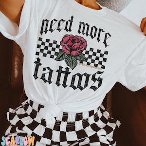 May include: White t-shirt with black checkered pattern and a red rose with green leaves. The text "need more tattoos" is written in black gothic font.