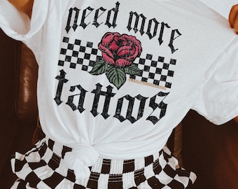 May include: White t-shirt with black checkered pattern and a red rose with green leaves. The text "need more tattoos" is written in black gothic font.