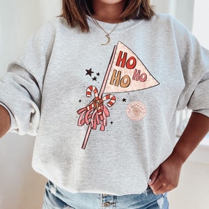 May include: A gray sweatshirt with a festive design featuring a pink and red pom-pom, candy canes, and a banner that reads "HO HO HO".