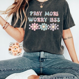 May include: A dark grey t-shirt with the text "Pray More Worry Less" in a retro font. The text is surrounded by colorful flowers.