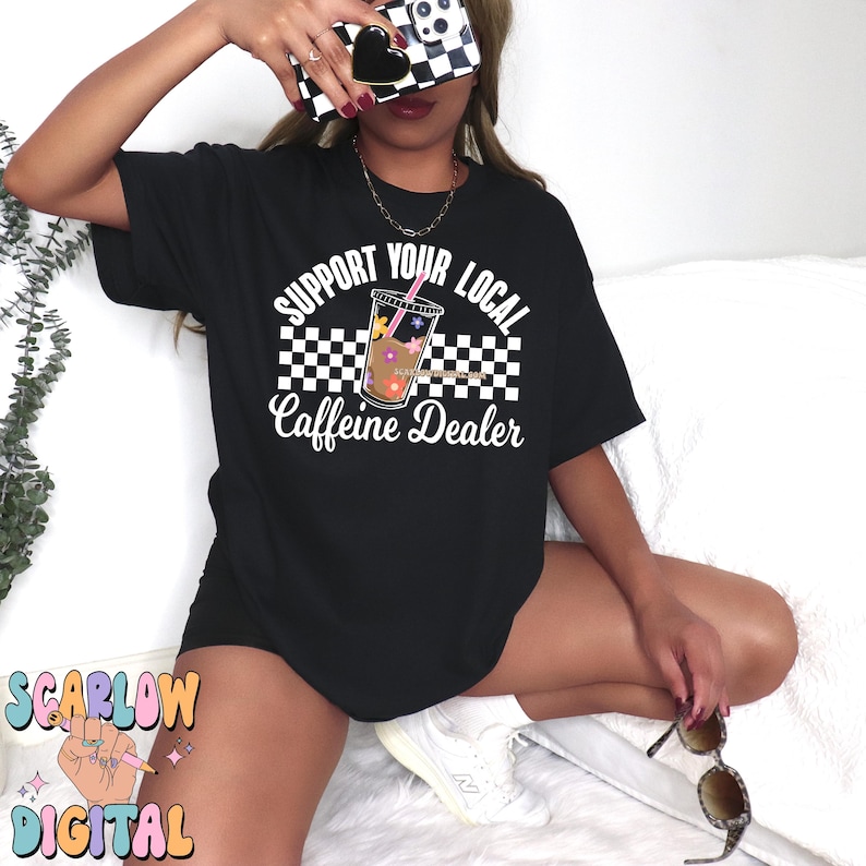 May include: Black t-shirt with a white graphic that says "Support Your Local Caffeine Dealer" with a checkered background and a cartoon image of a cup of iced coffee with a straw and flowers.