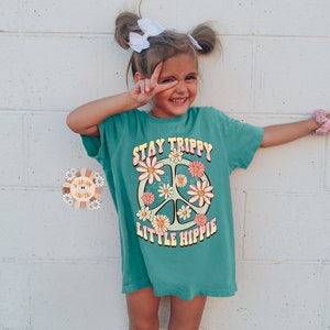 K&ouml;nnte beinhalten: Ein t&uuml;rkisfarbenes T-Shirt mit einem Peace-Zeichen-Motiv und dem Text "Stay Trippy Little Hippie".