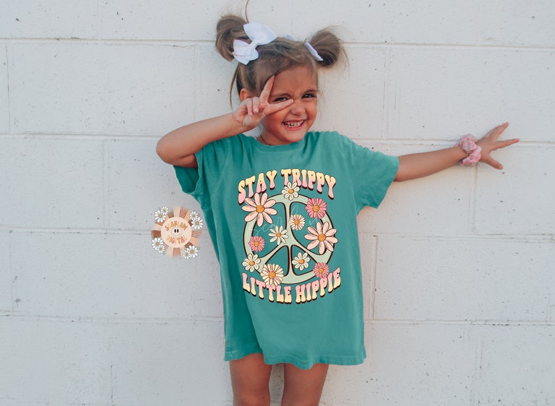 K&ouml;nnte beinhalten: Ein t&uuml;rkisfarbenes T-Shirt mit einem Peace-Zeichen-Motiv und dem Text "Stay Trippy Little Hippie".