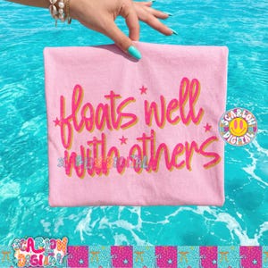 以下が含まれることがあります： ピンク色のTシャツ。重ねられたピンクと黄色のフォントで「floats well with others」というフレーズが書かれています。 ターコイズ色のマニキュアを塗った手でTシャツが持たれています。背景はターコイズ色の水のパターンです。