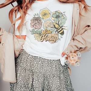 May include: White t-shirt with a graphic of a bee and flowers in a honeycomb design. The text "Sweet Like Honey" is written in a retro font.
