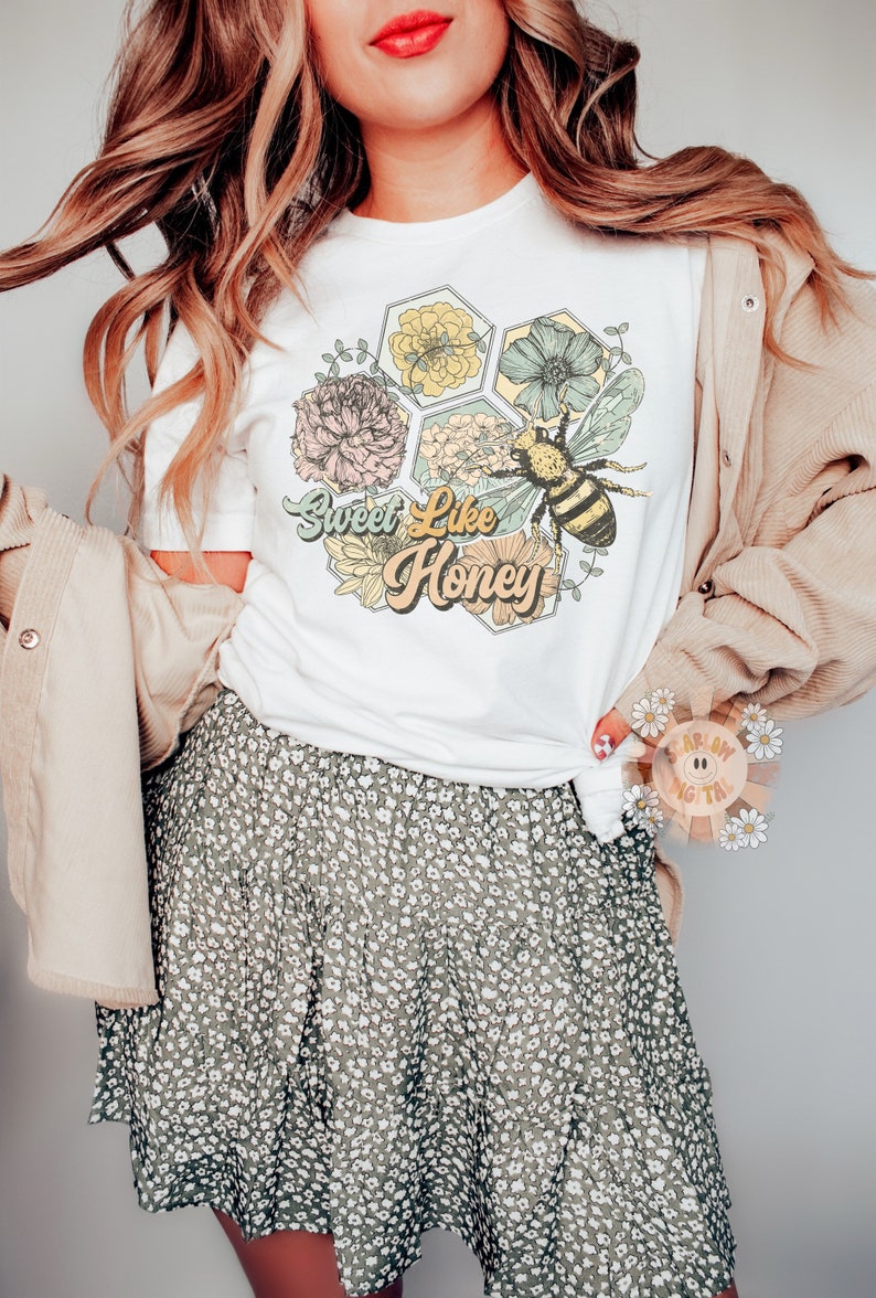 May include: White t-shirt with a graphic of a bee and flowers in a honeycomb design. The text "Sweet Like Honey" is written in a retro font.