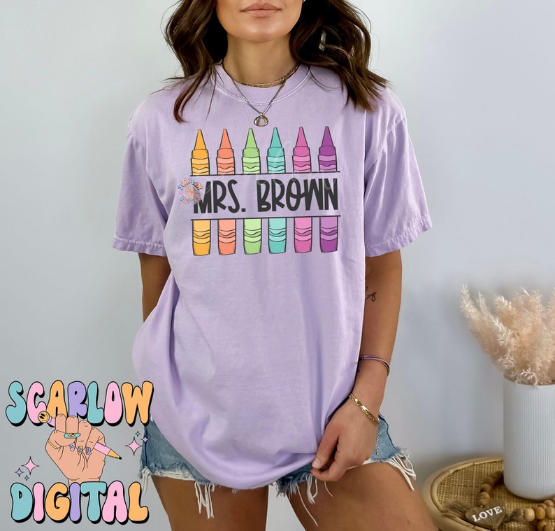 以下が含まれることがあります： 紫色のTシャツに、カラフルなクレヨンが並んだデザインと「MRS. BROWN」の文字がプリントされています。
