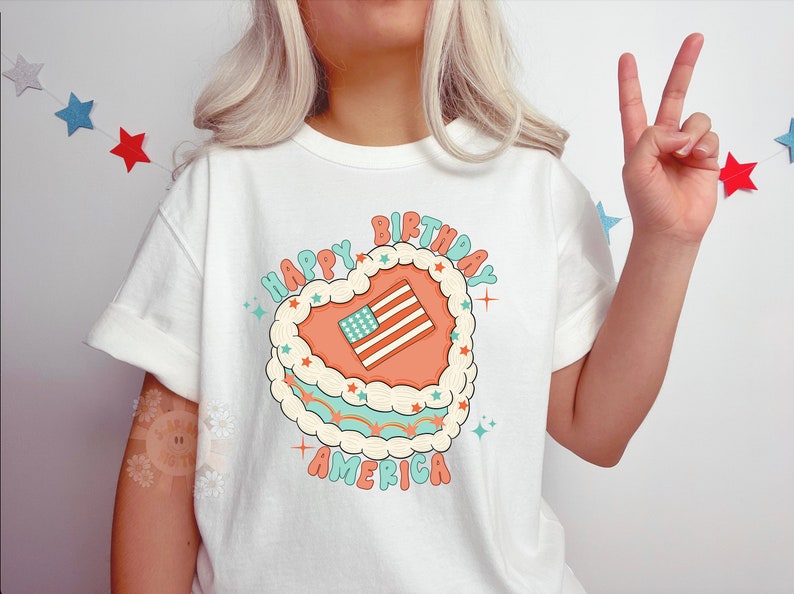 K&ouml;nnte beinhalten: Wei&szlig;es T-Shirt mit einem Aufdruck eines herzf&ouml;rmigen Kuchens mit einer amerikanischen Flagge darauf. Der Text "Happy Birthday America" ist auf dem T-Shirt aufgedruckt.