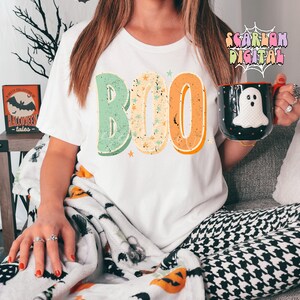 Peut inclure: Un t-shirt blanc avec le mot "BOO" imprim&eacute; dessus dans un style r&eacute;tro avec des couleurs vertes, orange et jaunes. Les lettres sont d&eacute;cor&eacute;es d'&eacute;toiles blanches. Le t-shirt est port&eacute; par une personne assise sur un canap&eacute; avec une couverture &agrave; motifs noirs et blancs.