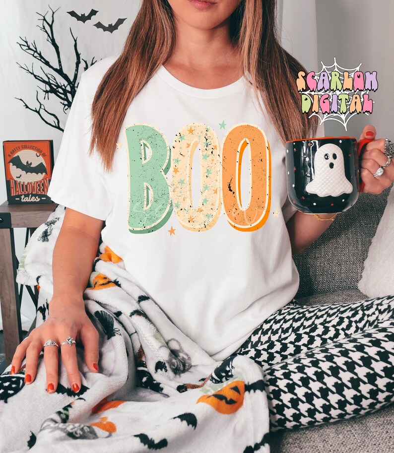 Peut inclure: Un t-shirt blanc avec le mot "BOO" imprim&eacute; dessus dans un style r&eacute;tro avec des couleurs vertes, orange et jaunes. Les lettres sont d&eacute;cor&eacute;es d'&eacute;toiles blanches. Le t-shirt est port&eacute; par une personne assise sur un canap&eacute; avec une couverture &agrave; motifs noirs et blancs.