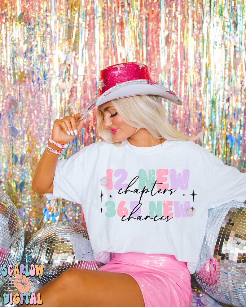 May include: A white t-shirt with a colorful graphic that says "12 new chapters + 36 new chances". The shirt is being worn by a person with blonde hair and a pink cowboy hat. The person is sitting in front of a silver disco ball.