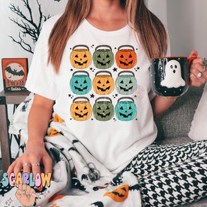 Puede incluir: Una camiseta blanca con un dise&ntilde;o de nueve cubos de calabaza de estilo vintage en varios tonos de naranja, verde y azul. Las calabazas tienen ojos, nariz y boca negros. El dise&ntilde;o est&aacute; centrado en la camiseta.