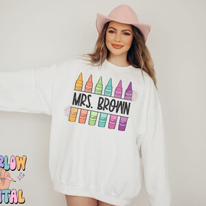 以下が含まれることがあります： カラフルなクレヨンと「MRS. BROWN」という黒いテキストのデザインが施された白いスウェットシャツです。