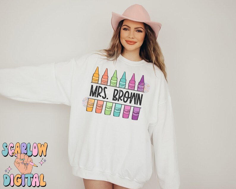 以下が含まれることがあります： カラフルなクレヨンと「MRS. BROWN」という黒いテキストのデザインが施された白いスウェットシャツです。