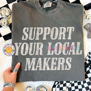 May include: A charcoal gray t-shirt with the text "SUPPORT YOUR LOCAL MAKERS" in a distressed white font. The shirt is displayed on a white and black checkered surface, with disco balls and other decorative items.