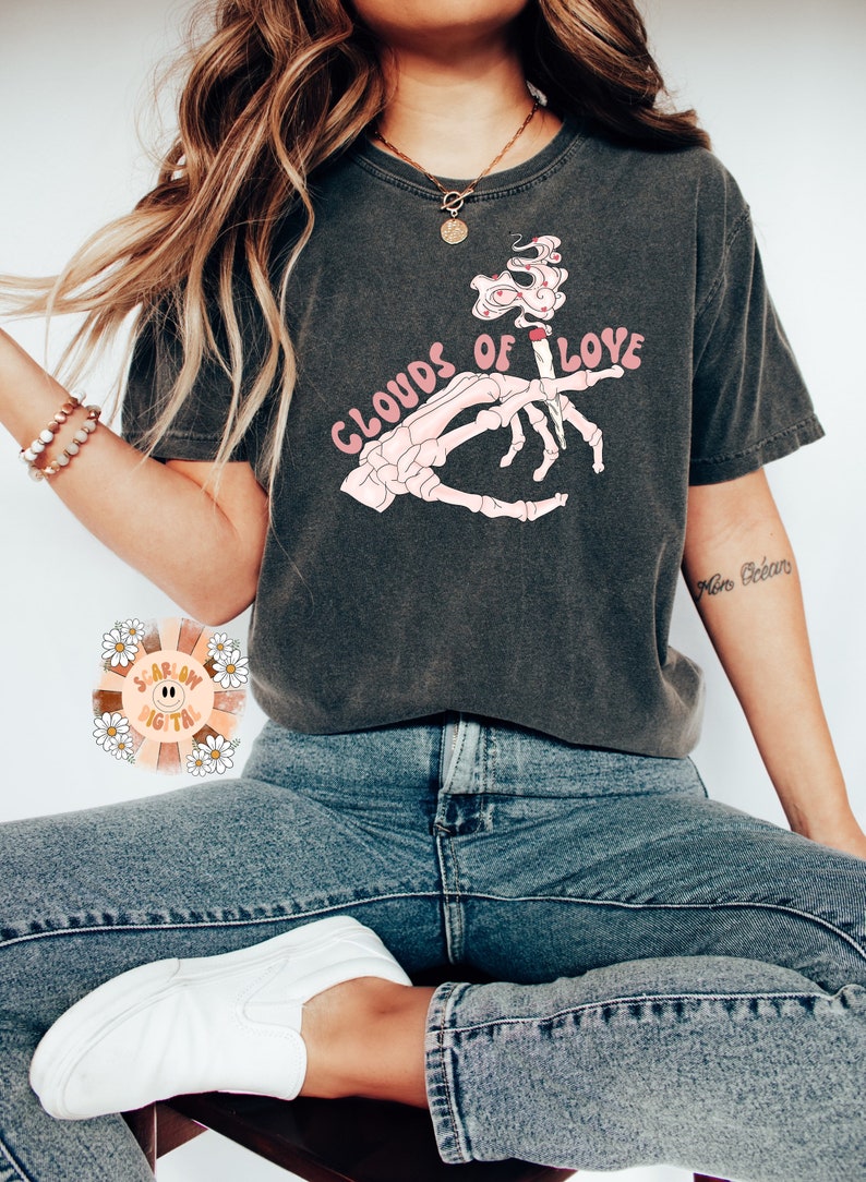 May include: Dark gray t-shirt with the text "CLOUDS OF LOVE" in pink. The graphic features a skeleton hand holding a lit cigarette with smoke shaped like hearts. The shirt is worn with blue jeans and white shoes.