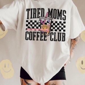 K&ouml;nnte beinhalten: Wei&szlig;es T-Shirt mit schwarz-wei&szlig;em Karomuster und dem Text "Tired Moms Coffee Club" in Schwarz. In der Mitte des Designs befindet sich eine Comic-Kaffeetasse mit einem rosa Strohhalm und bunten Verzierungen.