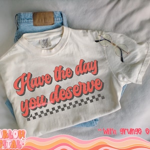 May include: White t-shirt with the text "Have the day you deserve" in red with a black and white checkered pattern underneath.