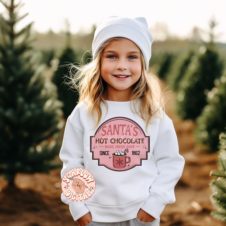 Op de afbeelding: Een wit sweatshirt met een roze en rode afbeelding waarop staat "Santa's Hot Chocolate - Made Fresh Daily - Since 1862". De afbeelding toont een mok met warme chocolademelk met marshmallows en een snoepstok.