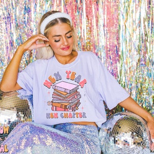 May include: A light purple t-shirt featuring a graphic of a stack of books and the text "New Year New Chapter".