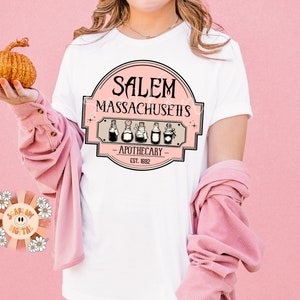 Pu&ograve; includere: Maglietta bianca con un design grafico rosa che dice "Salem Massachusetts - Apothecary - Est. 1832" con una fila di cinque bottiglie di pozioni.