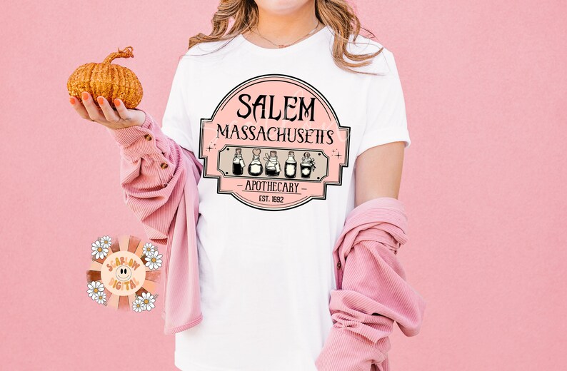 Pu&ograve; includere: Maglietta bianca con un design grafico rosa che dice "Salem Massachusetts - Apothecary - Est. 1832" con una fila di cinque bottiglie di pozioni.