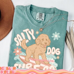 May include: A teal blue t-shirt with a graphic of a brown dog surfing on a surfboard. The text "Salty Dog Surf Co." is printed on the shirt. The shirt has a distressed look and is made of a soft, comfortable fabric.