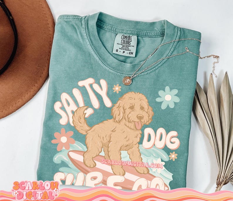 May include: A teal blue t-shirt with a graphic of a brown dog surfing on a surfboard. The text "Salty Dog Surf Co." is printed on the shirt. The shirt has a distressed look and is made of a soft, comfortable fabric.