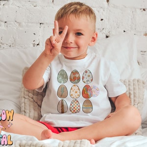 May include: A young child wearing a white t-shirt with a colorful Easter egg design. The eggs are decorated with various patterns, including stripes, polka dots, and geometric shapes.