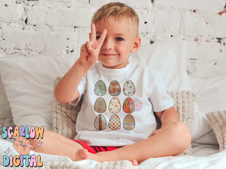 May include: A young child wearing a white t-shirt with a colorful Easter egg design. The eggs are decorated with various patterns, including stripes, polka dots, and geometric shapes.