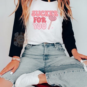 Może przedstawiać: Biało-czarna koszulka bejsbolowa z r&oacute;żowym i białym napisem "SUCKER FOR YOU" i grafiką r&oacute;żowego lizak w kształcie serca.