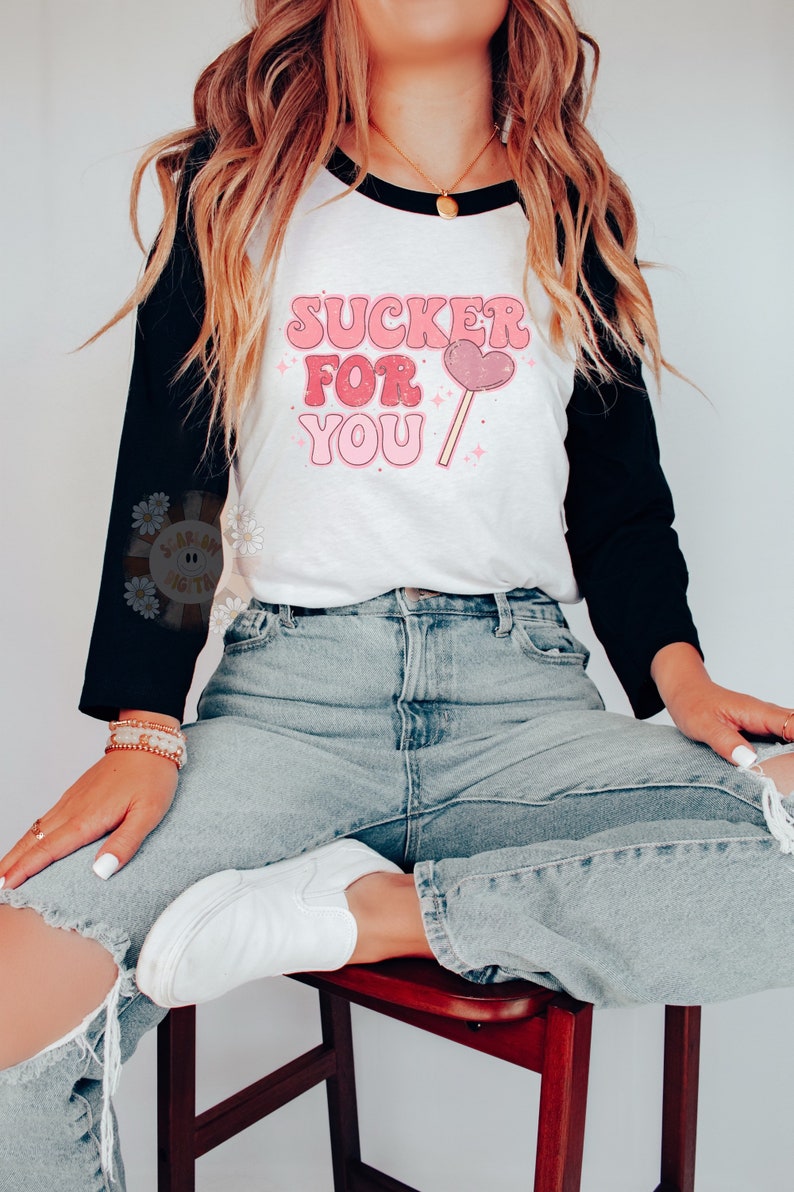 Może przedstawiać: Biało-czarna koszulka bejsbolowa z r&oacute;żowym i białym napisem "SUCKER FOR YOU" i grafiką r&oacute;żowego lizak w kształcie serca.