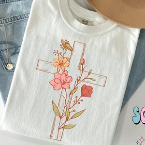 May include: White t-shirt with a floral design featuring a cross. The flowers are pink, orange, and yellow.