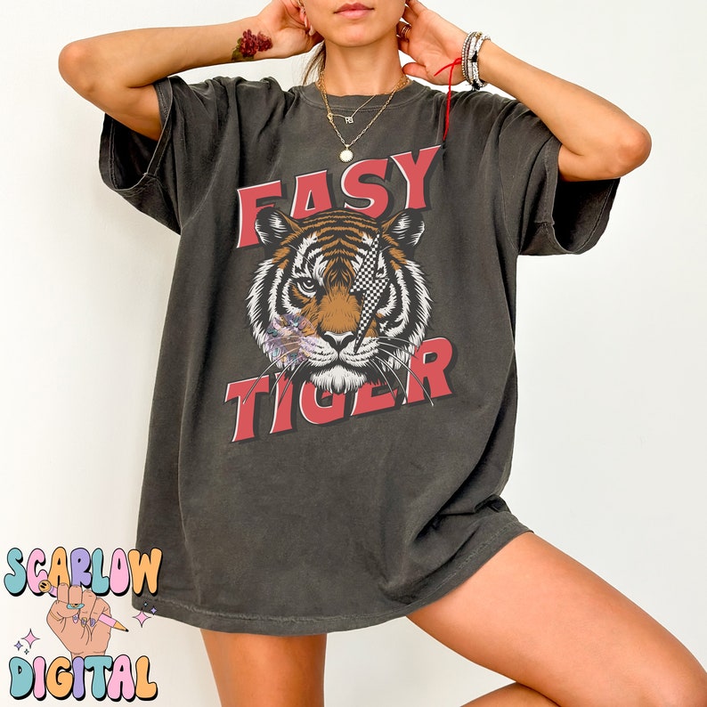 Puede incluir: Una camiseta de gran tama&ntilde;o de color gris oscuro con un estampado gr&aacute;fico de un tigre con un parche en el ojo a cuadros. El texto "EASY TIGER" est&aacute; impreso en rojo sobre el tigre.