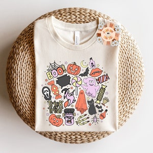 Peut inclure: Un t-shirt cr&egrave;me avec un motif color&eacute; sur le th&egrave;me d'Halloween. Le motif pr&eacute;sente une vari&eacute;t&eacute; d'&eacute;l&eacute;ments effrayants et amusants, notamment des fant&ocirc;mes, des citrouilles, des chauves-souris, des bonbons et les mots "Trick or Treat".