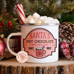 Op de afbeelding: Witte emaille mok met een rode en witte pepermuntstok die eruit steekt. De mok is gevuld met witte marshmallows en heeft een roze en wit label met de tekst "Santa's Hot Chocolate Made Fresh Daily Since 1862".