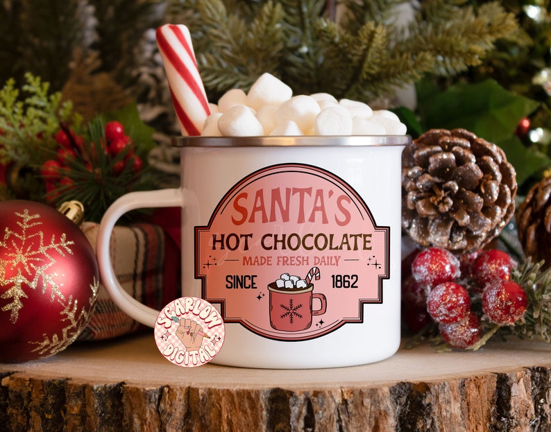 Op de afbeelding: Witte emaille mok met een rode en witte pepermuntstok die eruit steekt. De mok is gevuld met witte marshmallows en heeft een roze en wit label met de tekst "Santa's Hot Chocolate Made Fresh Daily Since 1862".
