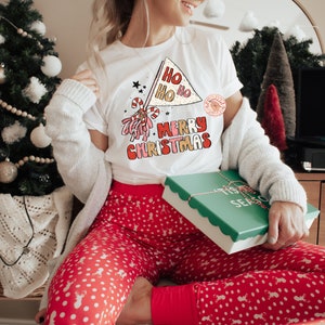 May include: A white t-shirt with a red, white, and gold graphic that says "Ho Ho Ho Merry Christmas". The person is wearing red and white polka dot pajamas and holding a green gift box with a white ribbon and the words "Tis the Season" on it.