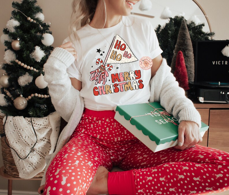 May include: A white t-shirt with a red, white, and gold graphic that says "Ho Ho Ho Merry Christmas". The person is wearing red and white polka dot pajamas and holding a green gift box with a white ribbon and the words "Tis the Season" on it.
