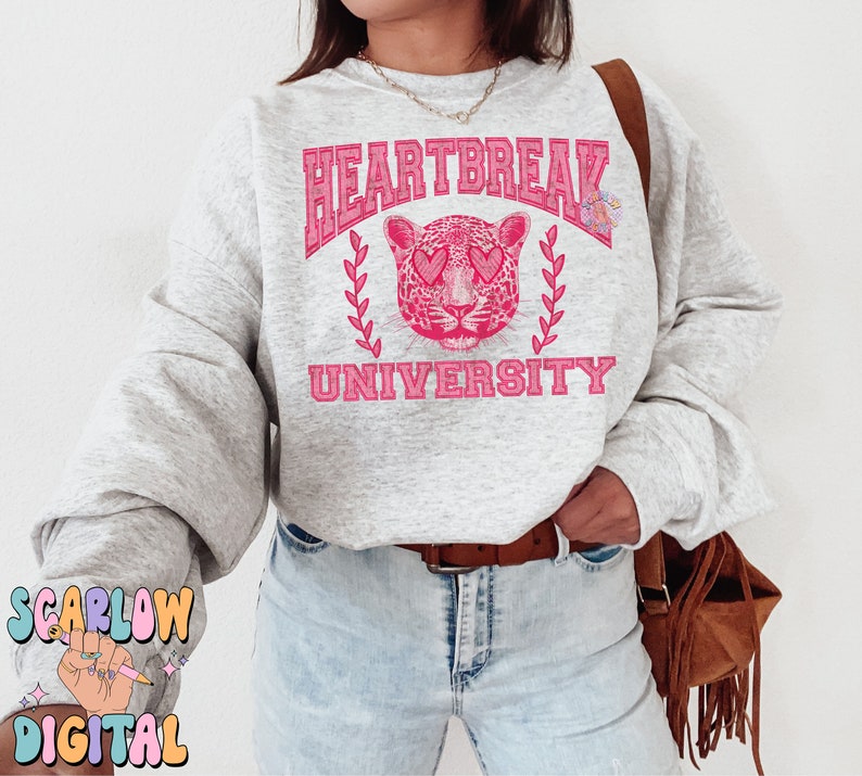 Pu&ograve; includere: Una felpa grigia con un design grafico rosa che presenta un leopardo con cuori per occhi e il testo "Heartbreak University".
