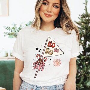 May include: White t-shirt with a pink and red pom-pom, candy cane, and a flag that says "HO HO HO" printed on it.