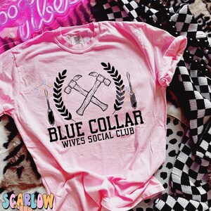 May include: Pink t-shirt with a black graphic of crossed hammers and a screwdriver with a wreath around them. The text "Blue Collar Wives Social Club" is below the graphic.
