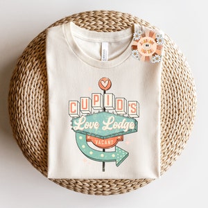 以下が含まれることがあります： ネオンサインのレトロなグラフィックがプリントされた白いTシャツ。サインには「キューピッドのラブロッジ」と表示され、「空室」のサインが付いています。サインは緑、青、オレンジで、白いドットが散りばめられています。
