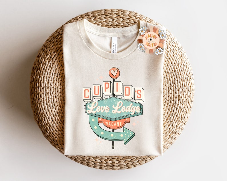 以下が含まれることがあります： ネオンサインのレトロなグラフィックがプリントされた白いTシャツ。サインには「キューピッドのラブロッジ」と表示され、「空室」のサインが付いています。サインは緑、青、オレンジで、白いドットが散りばめられています。
