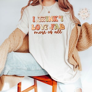 Op de afbeelding: Een wit T-shirt met de tekst "I think I love fall most of all" in kleurrijke, gepatroneerde letters. De letters zijn versierd met kleine bloemenmotieven.