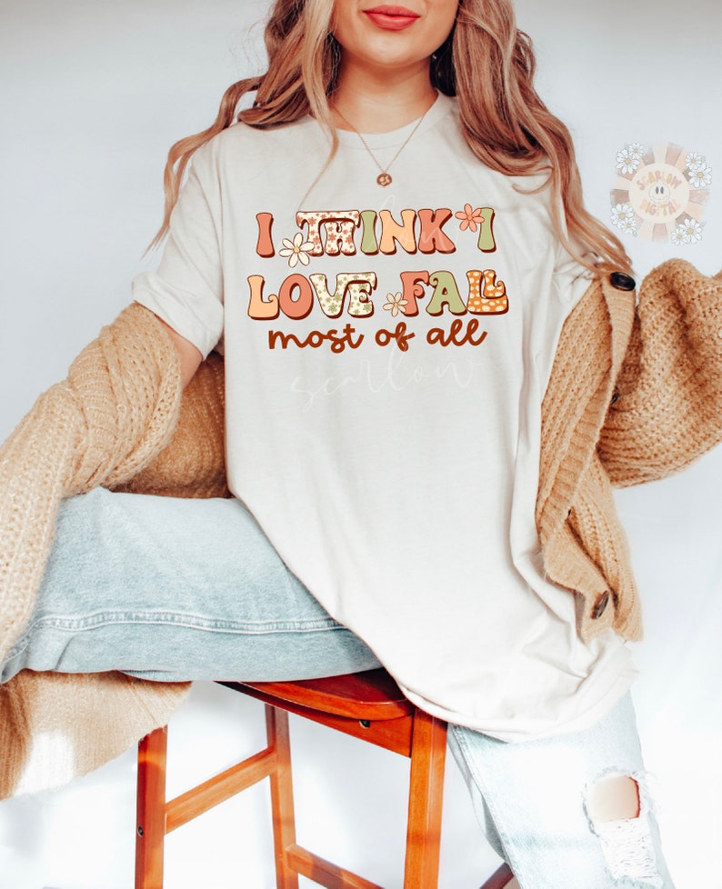 Op de afbeelding: Een wit T-shirt met de tekst "I think I love fall most of all" in kleurrijke, gepatroneerde letters. De letters zijn versierd met kleine bloemenmotieven.