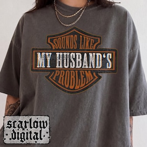 Puede incluir: Una camiseta de manga corta de color gris carbón con un diseño gráfico en forma de escudo con texto en naranja y blanco que dice "SOUNDS LIKE MY HUSBAND'S PROBLEM". La camiseta tiene cuello redondo y un corte holgado y relajado.