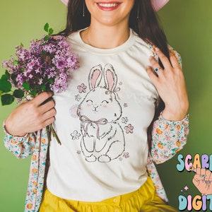 May include: White t-shirt with a pink and white bunny rabbit graphic with flowers and a pink bow. The bunny is holding a pink flower.
