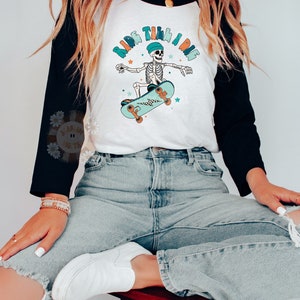 K&ouml;nnte beinhalten: Ein wei&szlig;es und schwarzes Baseball-T-Shirt mit einem Skelett, das Skateboard f&auml;hrt, und dem Text "Ride Til I Die" in einer bunten Schrift.