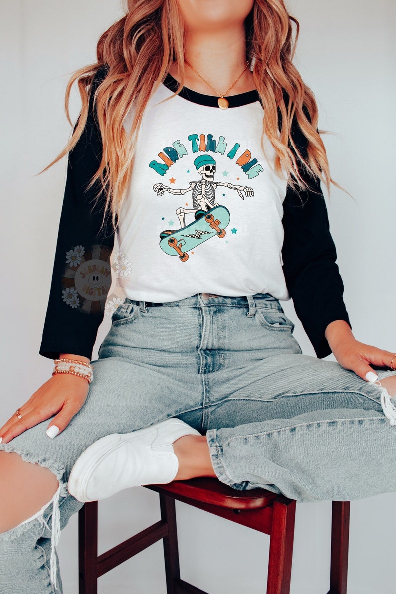 K&ouml;nnte beinhalten: Ein wei&szlig;es und schwarzes Baseball-T-Shirt mit einem Skelett, das Skateboard f&auml;hrt, und dem Text "Ride Til I Die" in einer bunten Schrift.